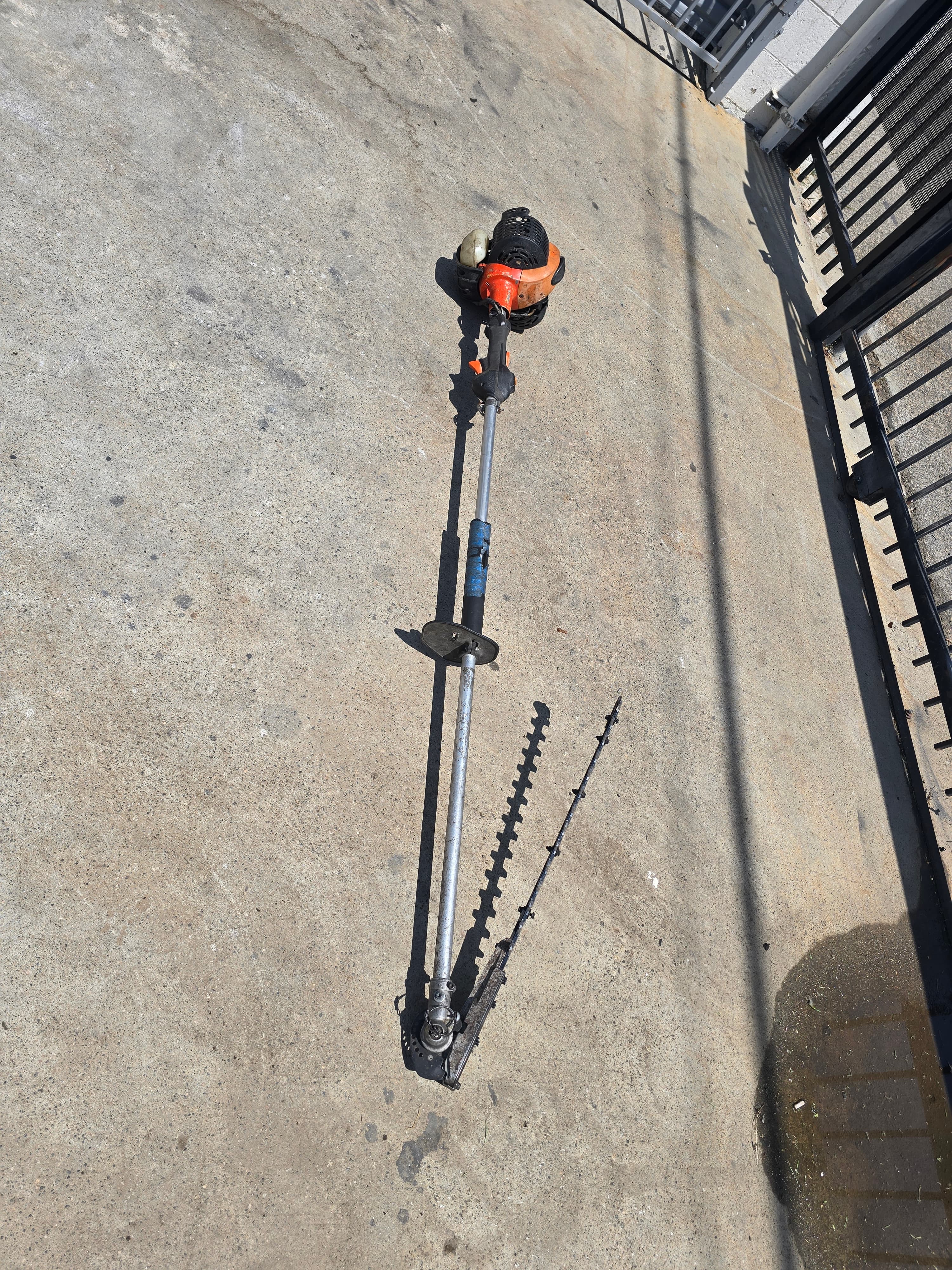 Orange gas-powered hedge trimmer with a broken blade attachment lying on a concrete surface.