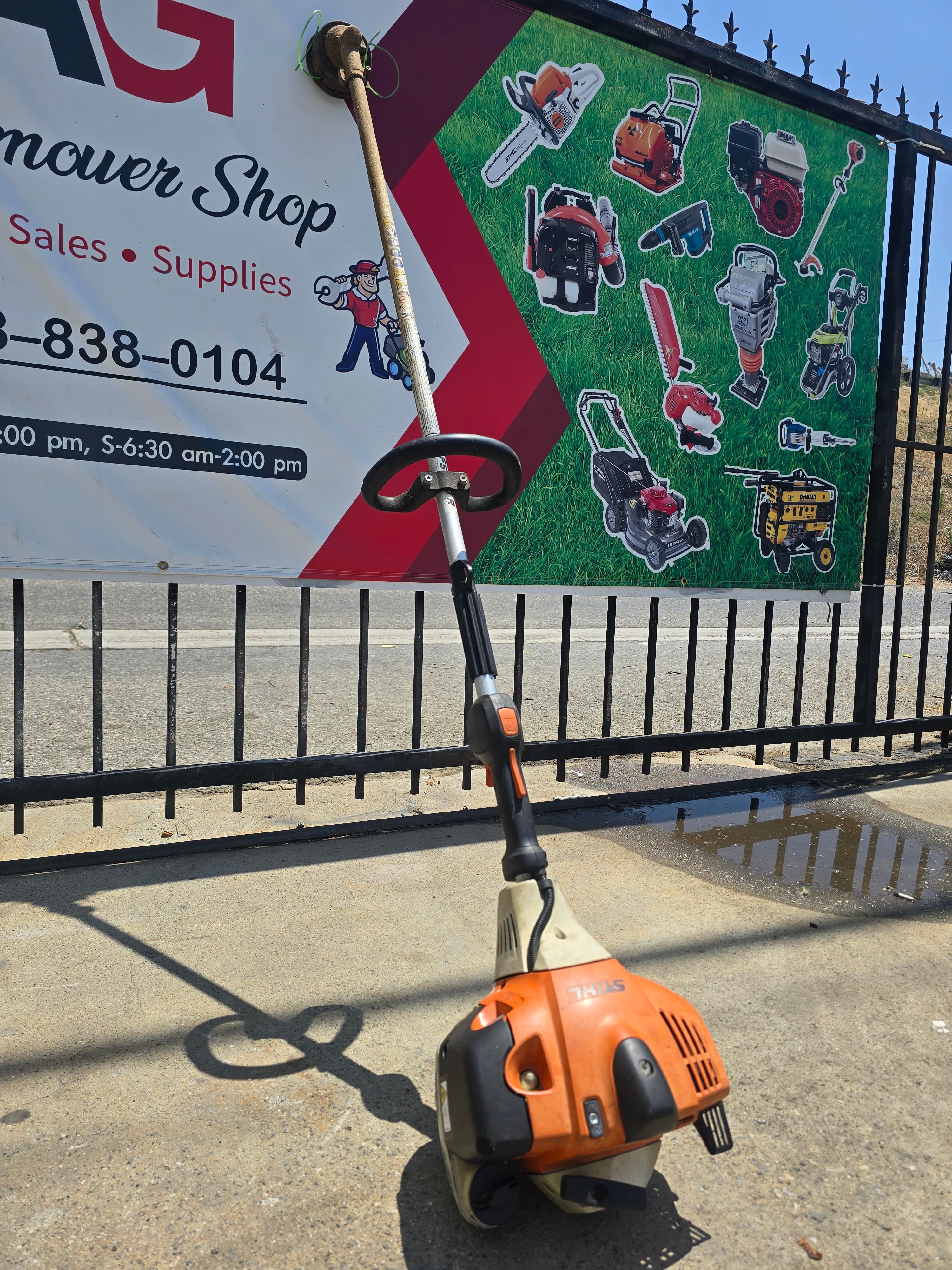 Orange Stihl string trimmer leaning against a black fence with a mower shop banner.