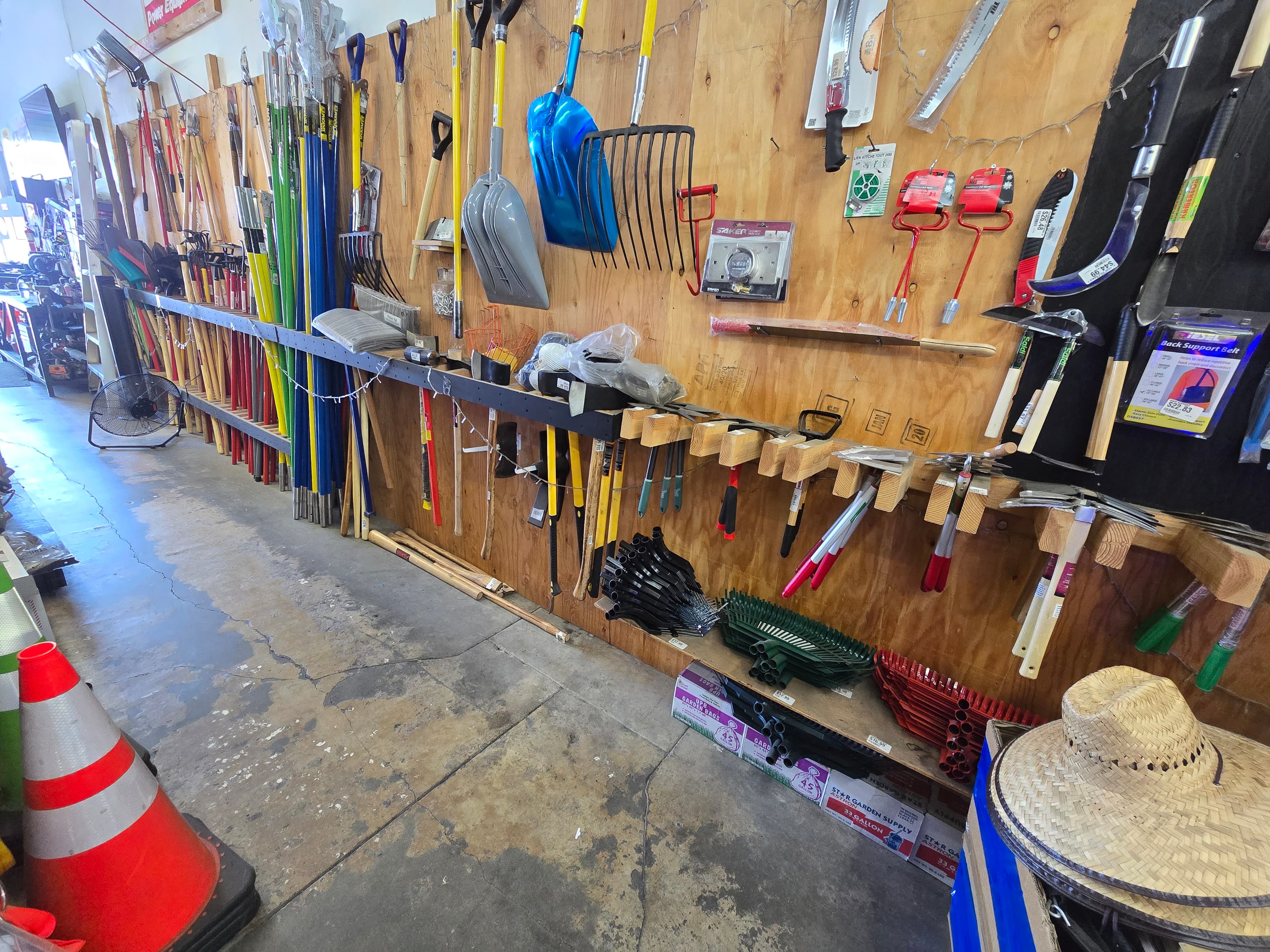 Assorted shovels, rakes, and hand tools displayed on a plywood wall in a supply store.